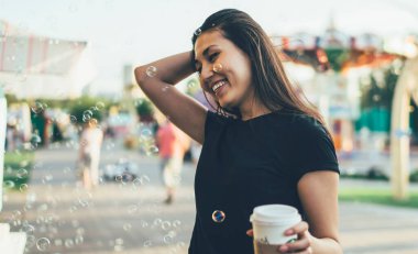 Gündelik giyinmiş mutlu Asyalı genç kadın elinde kahve tutarak şehir festivalinde tatil yaparken havada sabun köpüklerinin tadını çıkarıyor. Hafta sonları parkta eğlenen neşeli hippi kız.