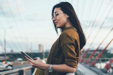 Genç Asyalı moda göz giyimli kadın sosyal ağlardan haberleri okuyor akıllı telefondan köprü kurulumu üzerinde dinleniyor, seyahat sırasında cep telefonunu paylaşan kadın seyahat blogcusu