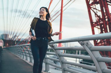 Keyifli Asyalı modaya uygun giyinmiş hippi kız şehir planlama köprüsünde yürümekten keyif alıyor, açık havada gezen gülümseyen kız öğrenci yolculuk sırasında modern tasarım şehir mimarisine bakıyor.