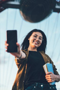 Moda gözlüklü ve giyimli neşeli Asyalı yeniyetme kızın modern akıllı telefonu boş ekran fotokopi alanı ile bulanık bir tonajda gösteren şehir ortamında duruş portresi