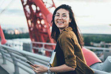 Gözlüklü neşeli Asyalı uluslararası öğrencinin portresi köprü üzerinde cep telefonu tutarak blog yazmak için zaman harcıyor, gülümseyen kadın gezgin arama yeri aramaktan memnun.