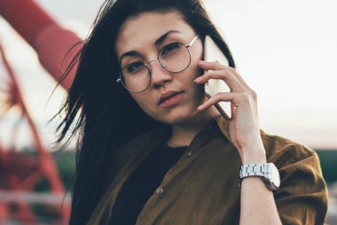 Gözlüklü çekici bir genç kadın yolcunun yarım boy portresi akıllı telefon görüşmesi yapıyor, yurtdışına seyahat ederken cep telefonuyla konuşan dalgın hippi kız kameraya bakıyor.