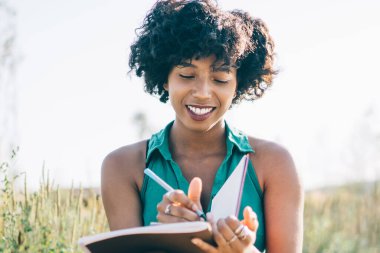 Gülümseyen, siyah saçlı, kıvırcık saçlı, planlamacıyla oturan, açık havada fikirlere dikkat çeken, mutlu, Afro-Amerikan hipster kız blog için yayın metni yazan, yedek kulübesinde eğlenen.