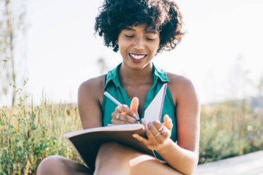 Gülümseyen, siyah saçlı, kıvırcık saçlı, planlamacıyla oturan, açık havada fikirlere dikkat çeken, mutlu, Afro-Amerikan hipster kız blog için yayın metni yazan, yedek kulübesinde eğlenen.