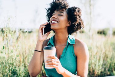 Neşeli Afrikalı Amerikalı hippi kız cep telefonuyla konuşuyor. Gülüyor ve kahve içiyor. Güneşli bir günde dışarı çıkıp eğleniyor.