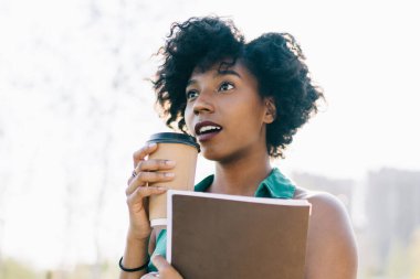 Çekici, siyah tenli, kıvırcık saçlı bir kadın açık havada kahve eşliğinde duran bir şeye hayran kalmış güzel, Afro-Amerikan hippi kız dışarıda sürpriz bir şekilde vakit geçirdiğini ifade ediyor.