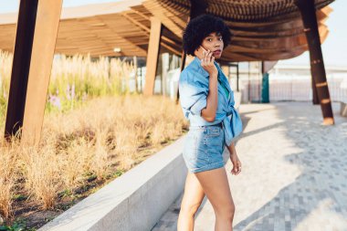 Çekici moda, koyu tenli hippi kız cep telefonuyla konuşuyor, şehir parkında yürüyor, genç Afrikalı Amerikalı kadın yaz güneşli bir günde şehri ziyaret ederken arama yapıyor.