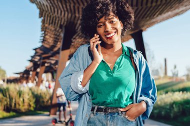 Kıvırcık saçlı, cep telefonuyla konuşan neşeli hippi kızın yarım boy portresi. Mutlu Afro-Amerikan kadını, aramalar için gümrük vergileriyle tatmin oluyor.