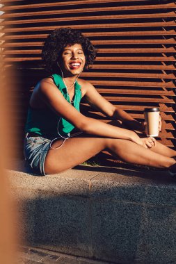 Neşeli Afrikalı Amerikalı hippi kızın portresi kıvırcık saçlı eğlendirici radyo çalma listesini akıllı telefondan dinleyen neşeli esmer tenli kadının güneşli bir günde gülüşü.