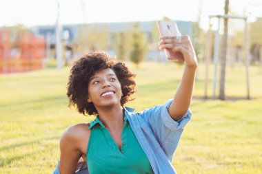 Çekici koyu tenli kadın blogcu modern akıllı telefon kamerasıyla sosyal ağları paylaşmak için selfie çekiyor. Çekici Afrikalı Amerikalı etkileyicisi boş zamanlarında video yapmak için cep telefonu kullanıyor.