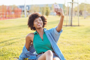 Çekici koyu tenli kadın blogcu modern akıllı telefon kamerasıyla sosyal ağları paylaşmak için selfie çekiyor. Çekici Afrikalı Amerikalı etkileyicisi boş zamanlarında video yapmak için cep telefonu kullanıyor.