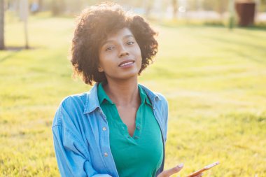 Siyah saçlı, yeşil çimenlerde oturan çekici bir kadının portresi. Cep telefonunu blog yazarken kullanıyor. Çekici Afrikalı Amerikalı hippi kız parkta elinde akıllı telefon ile kameraya bakıyor.