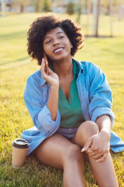 Yeşil çimenlerde oturan çekici hippi kız internet üzerinden akıllı telefon görüşmesi yapıyor koyu tenli kadın milenyum boyunca parkta cep telefonuyla konuşuyor.