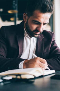 Düşünceli, beyaz sakallı bir adam not defteri planlama programına yazıyor. Ofisinde oturuyor, resmi giyinmiş düşünceli bir erkek girişimci iş süreci finans stratejilerini düzenleyen notlar yazıyor.