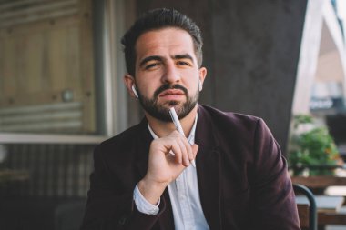 Resmi olarak giyinmiş ciddi bir hipster 'ın portresi kameraya bakıp kafeterya terasında oturup hayal ürünü beyaz iş adamının çalışma saatlerini not alarak yayınlama fikri üzerine kafa yoruyor.