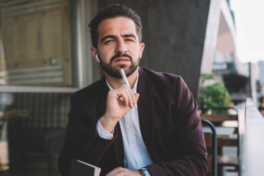 Resmi olarak giyinmiş ciddi bir hipster 'ın portresi kameraya bakıp kafeterya terasında oturup hayal ürünü beyaz iş adamının çalışma saatlerini not alarak yayınlama fikri üzerine kafa yoruyor.