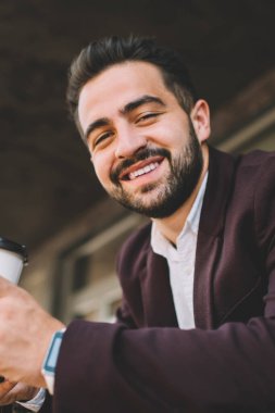 Resmi kıyafetli mutlu sakallı bir adam molada dinlenirken kameraya bakıyor, yakışıklı sakallı genç bir menajerin portresi başarılı kariyerinden memnun kalmış bir şekilde kahve tutuyor.