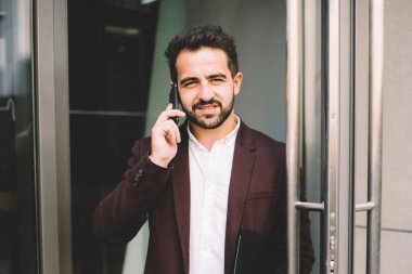 Resmi ceketli neşeli beyaz erkek binanın kapısında durmuş cep telefonuyla konuşuyor, zengin iş adamı gülümsüyor akıllı telefonla konuşuyor emlak binasına kamerayla bakıyor.