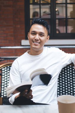 Neşeli modern Asyalı erkek okuyucu, kırmızı tuğla duvarlı kafede otururken komik bir kitap ve kahveyle vakit geçiriyor.