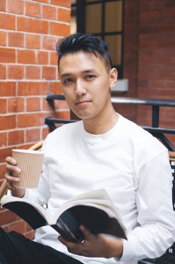 Masada oturup kafede dinlenirken beyaz gömlekli, kahve içen ve en sevdiği kitabı okuyan ciddi bir Asyalı erkek.