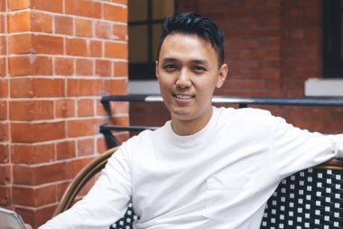 Tatmin olmuş Asyalı beyaz ceketli bir erkek öğrenci rahat açık hava kafeteryasında içki içerken cep telefonu kullanıyor ve kameraya bakıyor.