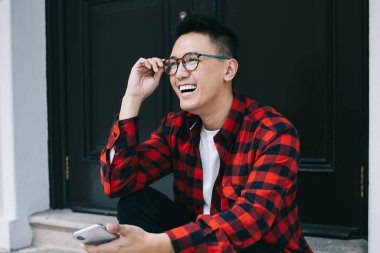 Şen şakrak genç, kareli tişörtlü, gözlüklü, akıllı telefon kullanırken ve bina kapısında merdivenlerde otururken gülen ve gözlüğe dokunan etnik bir adam.