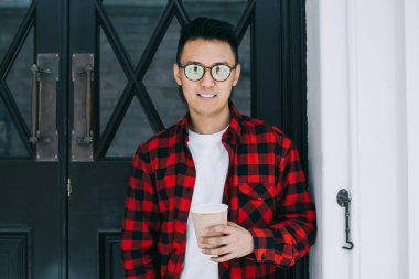 Gözlüklü ve kırmızı tişörtlü neşeli Asyalı adam elinde yemek fincanı tutarken kameraya bakıyor ve binanın kapısında dikiliyordu.