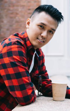 Kırmızı tişörtlü neşeli Asyalı bir erkek kameraya bakıyor, cep telefonunu tutuyor ve balkona yaslanıyor.