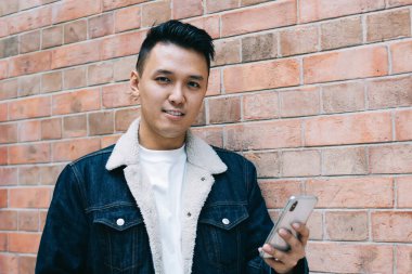 Tuğla duvara yaslanarak kameraya bakmak için kahveyle yürürken akıllı telefona göz atan kendinden emin Asyalı genç adam.