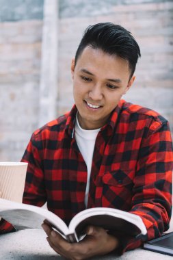Neşeli genç etnik erkek öğrenci elinde içecek ve kitap tutarken Old City Caddesi 'ndeki taş sınırında dikilip yaslanırken