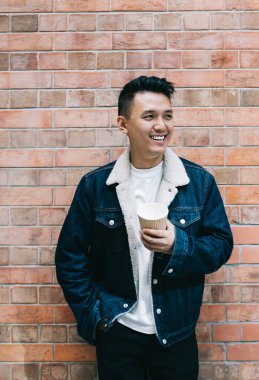 Asyalı iyimser erkek şehir duvarına yaslanıyor kahve içerken ve gülümseyerek uzaklara bakıyor.