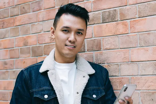 Tuğla duvara yaslanarak kameraya bakmak için kahveyle yürürken akıllı telefona göz atan kendinden emin Asyalı genç adam.