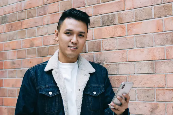 Tuğla duvara yaslanarak kameraya bakmak için kahveyle yürürken akıllı telefona göz atan kendinden emin Asyalı genç adam.