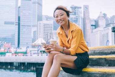 Neşeli gülümseyen yetişkin Asyalı kadın, rüzgarlı bir günde Hong Kong rıhtımının merdivenlerinde otururken elinde kahve fincanı ve akıllı telefonuyla gündelik kıyafetleriyle.