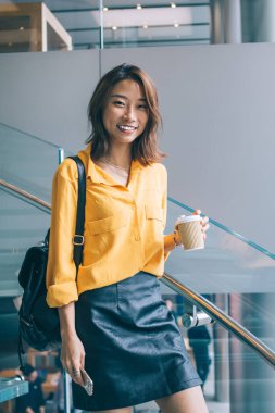 Halka açık bir yerde elinde kafein içeceğiyle poz veren neşeli Japon bayan öğrencinin yarım boy portresi. Pozitif Asyalı kadın gülümsüyor ve evde boş günün tadını çıkarıyor.