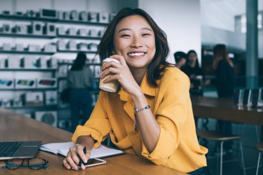 Gülümseyen Asyalı kız öğrenci, yemek arasında laptopuyla kahve içiyor ve Hong Kong 'daki elektronik mağazasında kameraya bakıyor.