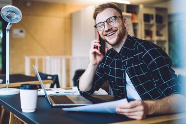 4G internete bağlı arkadaş canlısı iş arkadaşıyla mobil iletişimde gülen neşeli serbest yazar, görüş koruması için gözlüklü dişlek erkek blogcu cep telefonu sohbetinin keyfini çıkarıyor