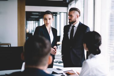 Bir grup ciddi yetişkin iş arkadaşı modern gökdelen ofisindeki büyük pencerenin yanındaki masada tartışıyorlar.