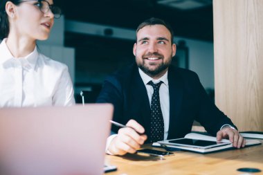 Resmi giysiler içinde, modern konferans odasındaki ahşap masada otururken tablet ve laptop kullanan özgüvenli yöneticiler bulanık arka planda sohbet ediyorlar.