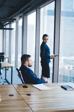 Resmi kıyafetli şık bir erkeğin cep telefonuyla konuşurken yan görünüşü açık ofis penceresinin yanında iş arkadaşlarıyla konuşuyor.