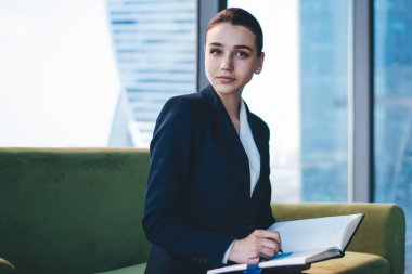 Ofis binasındaki kanepede oturup kameraya bakarken resmi takım elbiseli genç bir bayan not defterine notlar yazıyor.