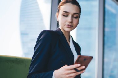 Genç konsantre iş kadını resmi takım elbise giyer. Kanepede elinde not defteriyle otururken modern akıllı telefona göz atar.