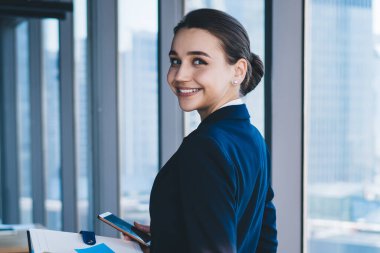 Modern gökdelenin penceresinin yanında durup kameraya bakarken iş ortağıyla cep telefonunda mesajlaşan neşeli kadın girişimcilerin yan görüntüsü