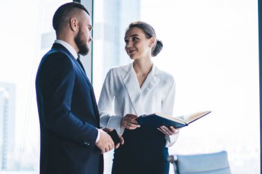 Resmi kıyafetleri içinde tanınmayan bir erkek CEO, modern ofis binasının penceresinde dururken elinde not defteriyle kadın muhasebeciyle konuşuyor.