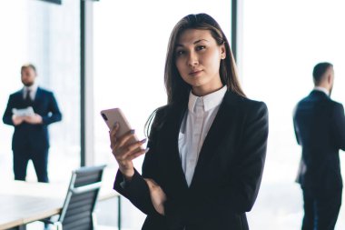 Ciddi Asyalı patron akıllı telefon kullanarak modern ofisin bulanık arka planında dururken kameraya bakıyor.