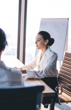 Aydınlık odada, modern iç mekanla rahat bir ofis koltuğunda otururken masa başında kitap okuyan meşgul kadın patronun yan görüntüsü.