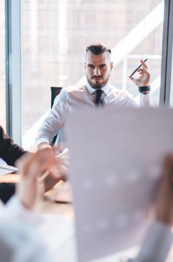 Modern şehir ofisindeki iş arkadaşlarıyla konferans masasında otururken resmi kıyafetli ciddi bir adam belgeyi tartışıyor ve dizüstü bilgisayarda geziniyor.