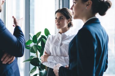 Takım elbiseli, sakallı bir yönetici. Geniş pencerenin yanında dikilip çalışırken ciddi genç kadınlarla konuşuyor.