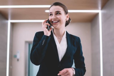 Neşeli ve neşeli bir ofis çalışanı. Parlak bir koridorda durmuş, akıllı telefondan konuşurken gülümsüyor ve gözlerini kaçırıyor.