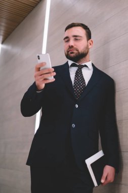 Saygıdeğer ve kendine güvenen resmi takım elbiseli erkek girişimci. Parlak odada duvara yaslanıyor ve sosyal ağlarda sohbet ederken defteri elinde tutuyor.